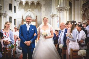 La mariee en robe blanche marchant dans l'allee centrale de l'eglise au bras de son pere devant les invites.