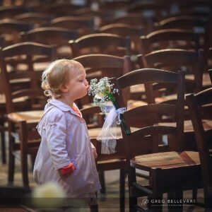 Une petite fille en manteau rose sentant délicatement une fleur blanche fixée à une chaise d'église.