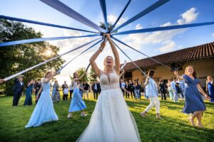 La mariee rit au centre d'un cercle de demoiselles d'honneur tenant des rubans bleus lors d'une animation de mariage en exterieur.