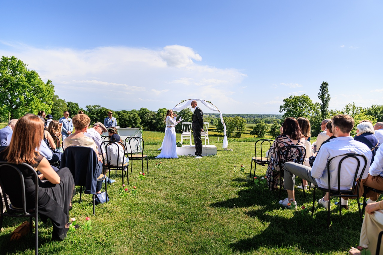 Vue d'ensemble d'une ceremonie de mariage en plein air avec les invites assis sur des chaises noires face aux maries sous une arche blanche.