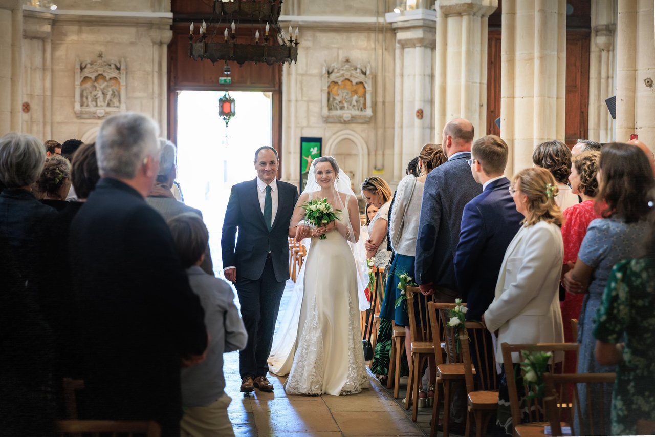 La mariée en robe blanche et voile avance dans l'allée centrale d'une église en pierre devant les invités.