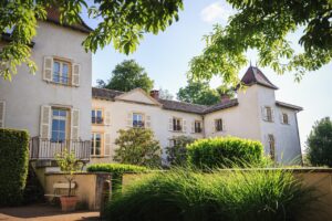 Façade en pierre claire du Château des Broyers entourée de verdure sous un ciel bleu.