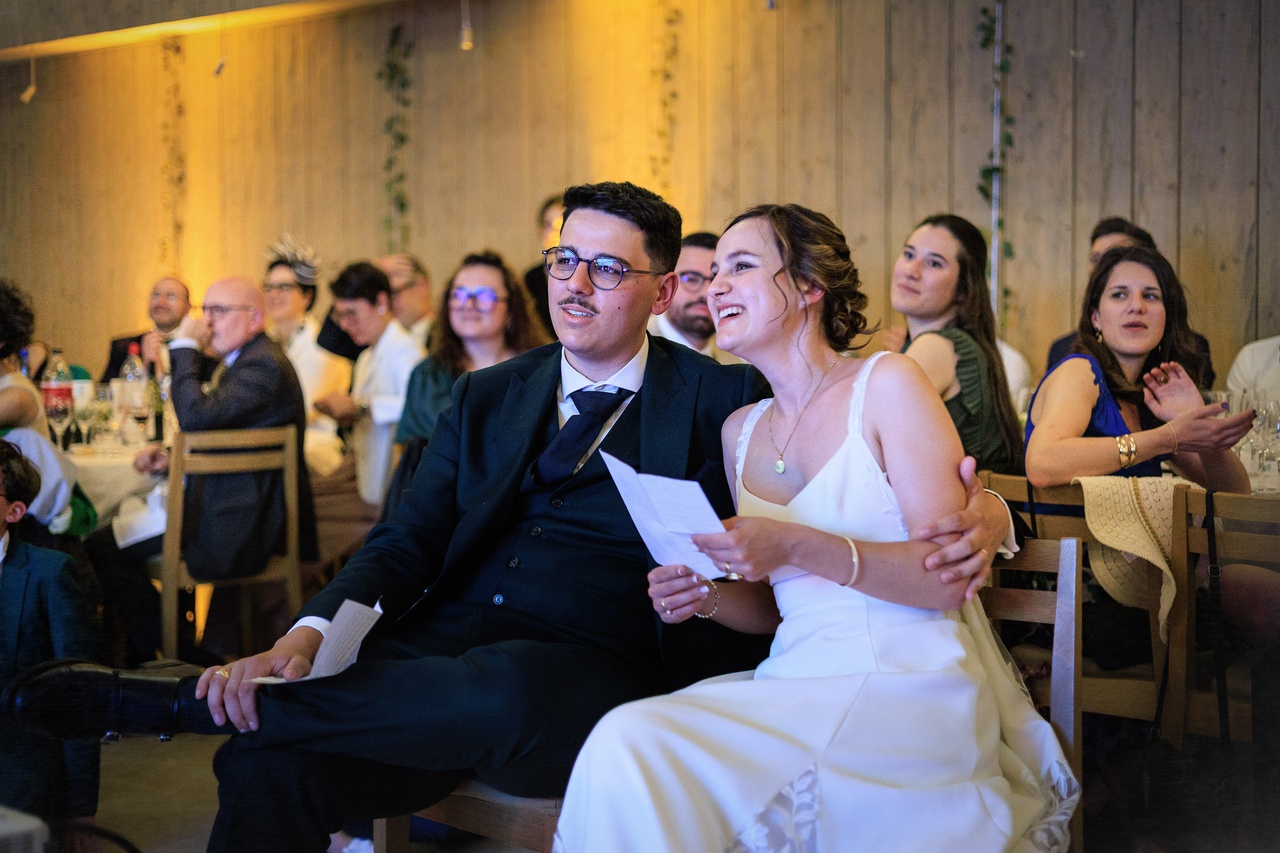 Les mariés assis à leur table écoutent un discours avec un regard ému et souriant.