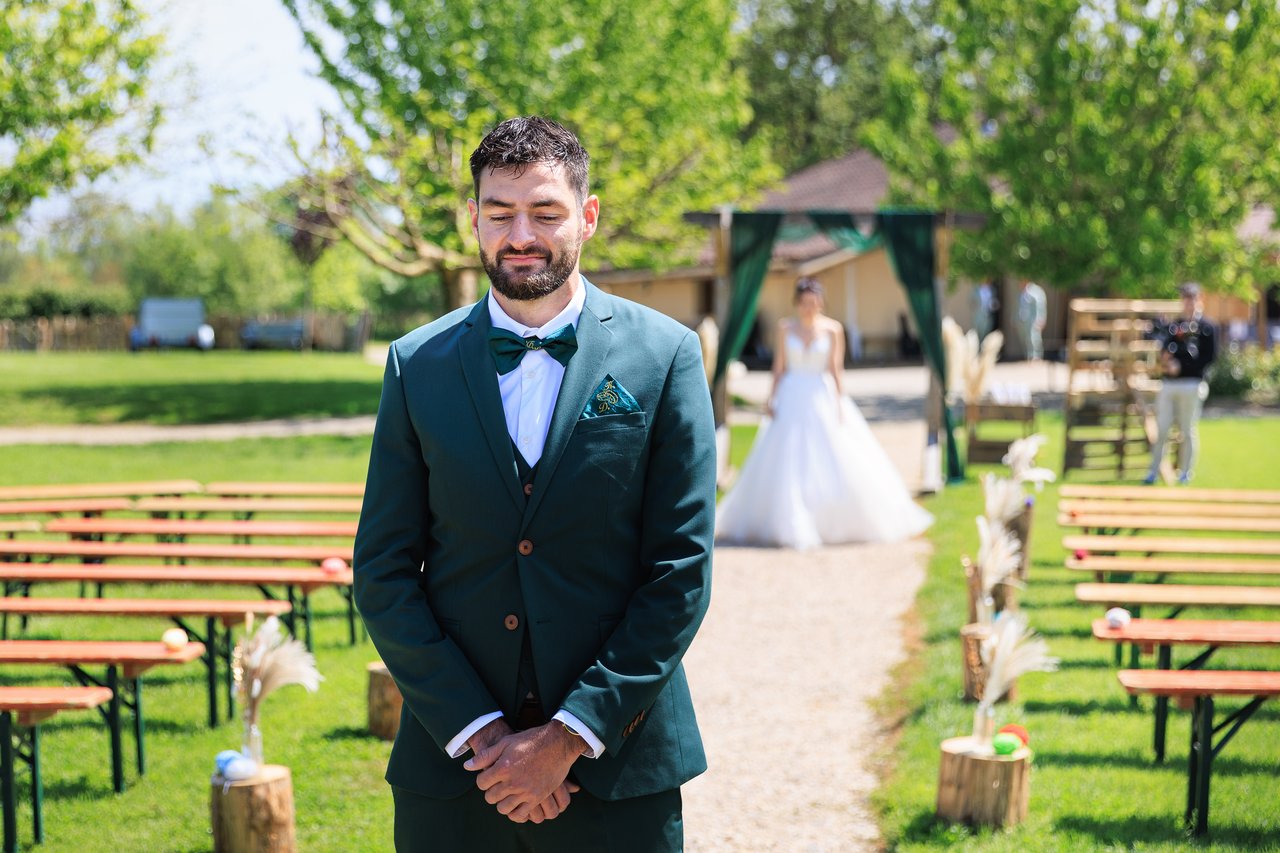 Le marie attend debout dans l'allee de la ceremonie avec la mariee au loin.