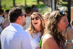 Portrait d'une invitee souriante portant des lunettes de soleil et une robe en dentelle blanche discutant avec un homme de dos.