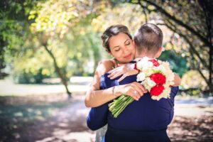 Mariee serrant son epoux dans ses bras avec un bouquet de roses rouges et blanches dans un parc arbore a Villefranche sur Saône