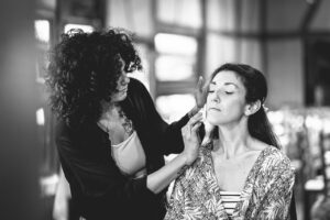 Photo en noir et blanc d'une maquilleuse appliquant du maquillage sur le visage de la mariee.
