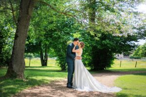 Portrait de couple des mariés s'embrassant debout dans une allée ombragée du jardin arboré du Château des Broyers.