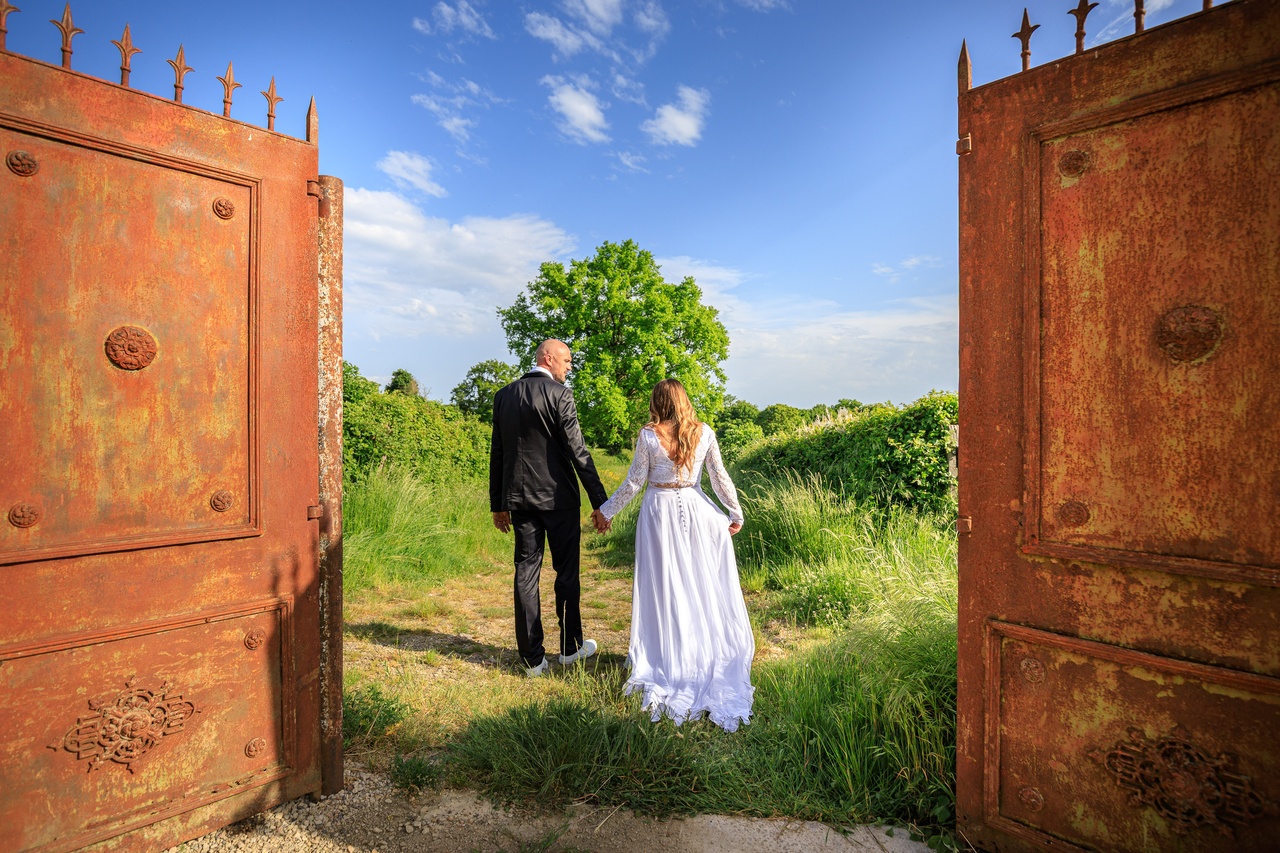 Les maries marchent main dans la main en s'eloignant de l'objectif a travers un grand portail en fer forge ouvert sur un champ.