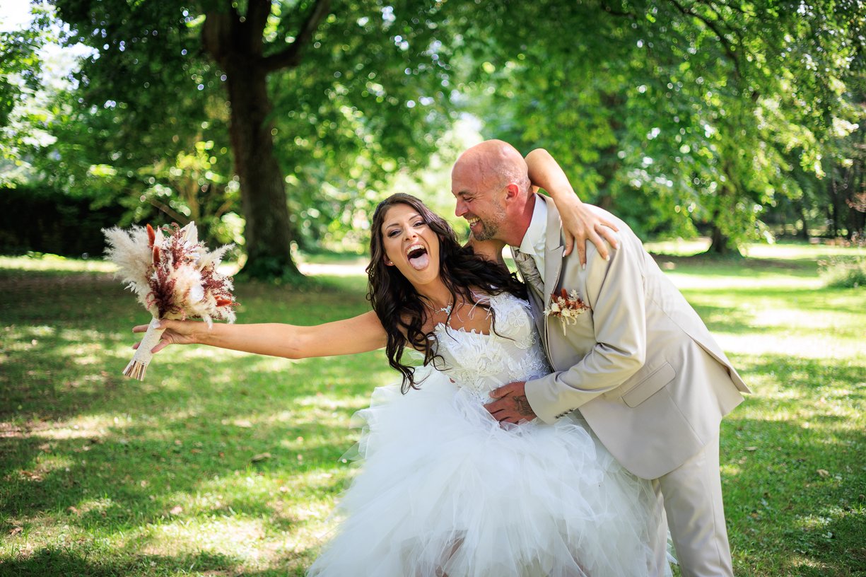 La mariée éclate de rire en tendant son bouquet, le marié la retient par la taille