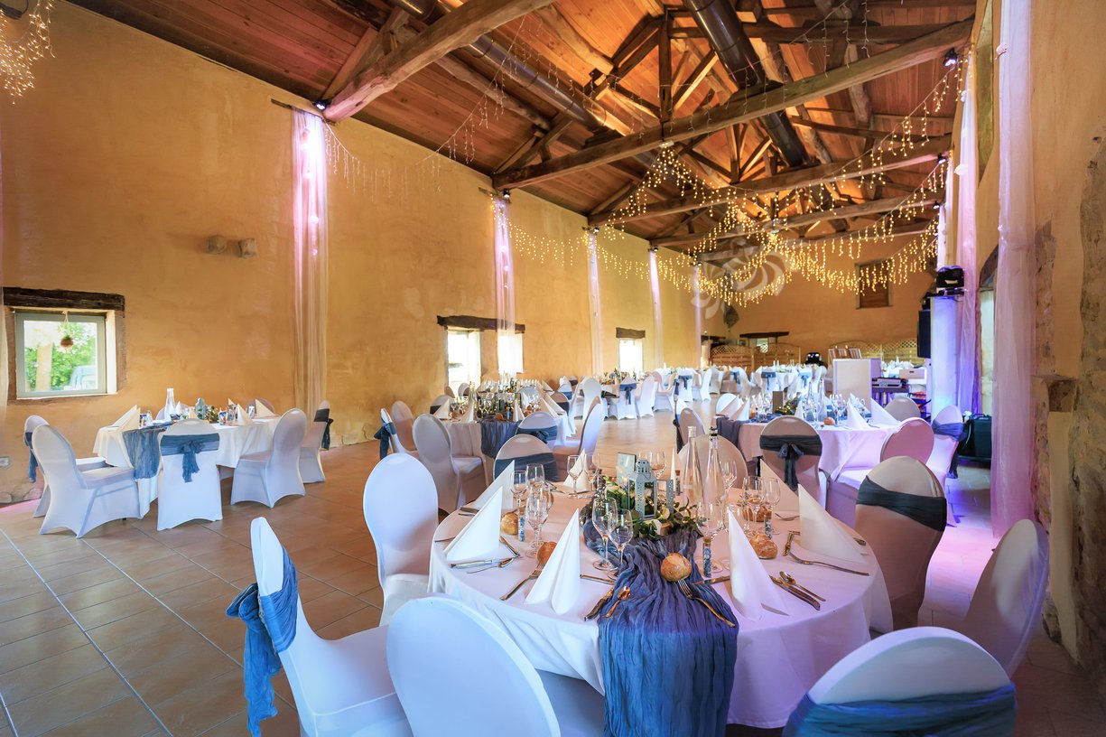 Salle de reception de la Ferme du Tremblay sous charpente, tables dressees en blanc et bleu, guirlandes lumineuses