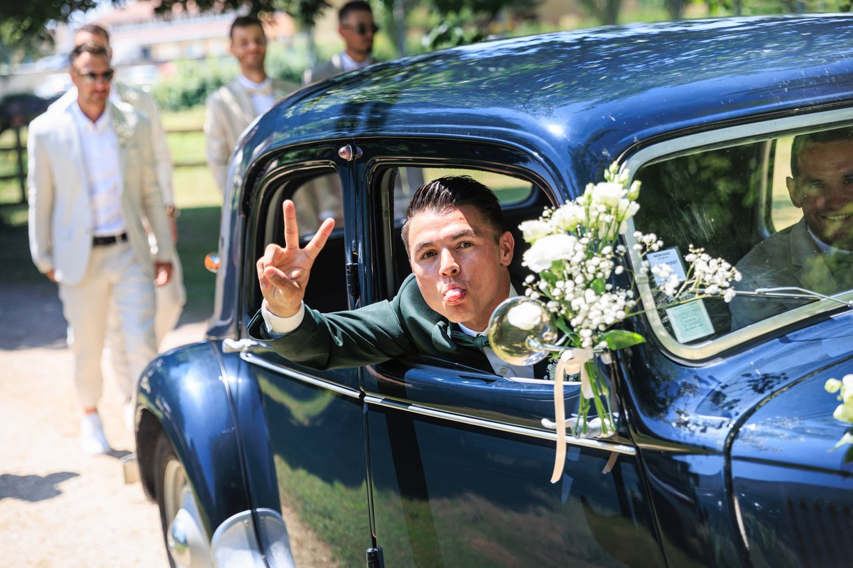 Dimitri passe la tete par la vitre d'une voiture ancienne et fait un signe de victoire, bouquet accroche a la portiere