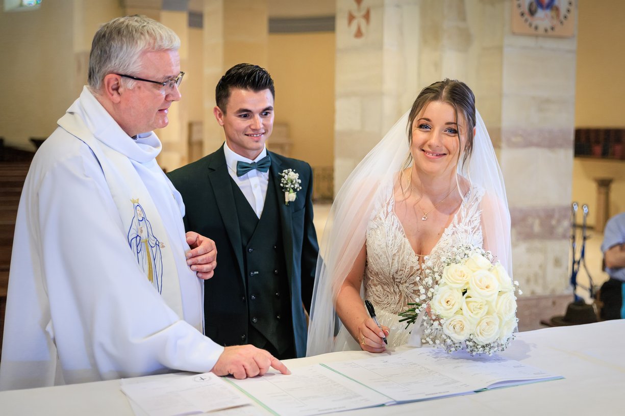 Camille et Dimitri signent les registres de mariage, le pretre a leurs cotes, tous les trois souriants